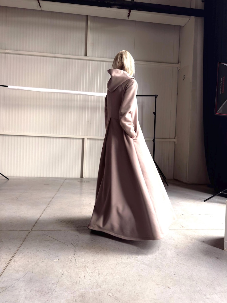 May include: A long, flowing, dusty rose-colored coat with a hood. The coat is floor-length and has a simple, elegant design. The model is standing in a studio setting, with a neutral background.