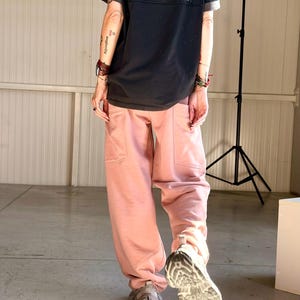 May include: A person wearing a black t-shirt with the text "LOW PLUNGE 0.79 AUDITORIUM 5th Edition" and pink sweatpants. The person is also wearing a blue denim cap and gray sneakers.