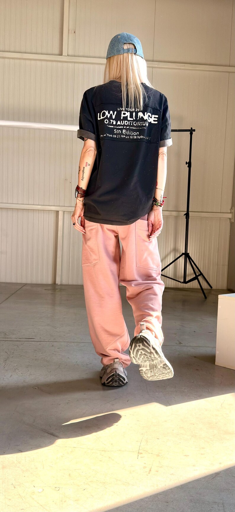 May include: A person wearing a black t-shirt with the text "LOW PLUNGE 0.79 AUDITORIUM 5th Edition" and pink sweatpants. The person is also wearing a blue denim cap and gray sneakers.