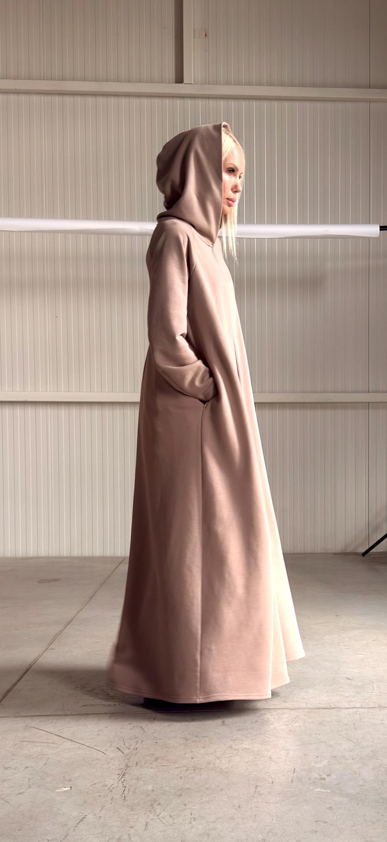 May include: A long, hooded, taupe-colored coat with pockets. The coat is floor-length and made of a soft material. The hood is large and drapes over the shoulders. A stylish and comfortable outerwear garment.