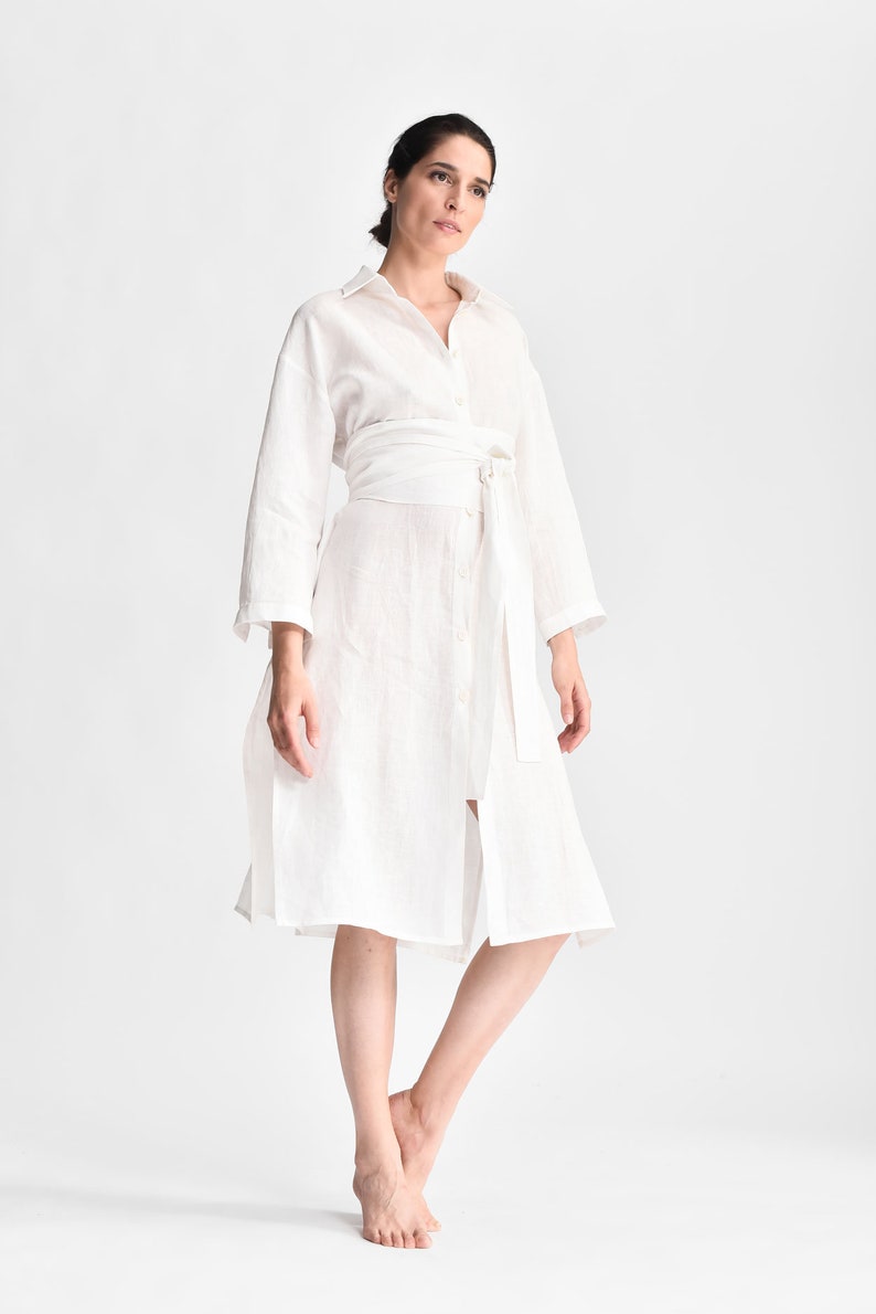 May include: A white linen shirt dress with a self-tie belt. The dress has a long sleeve and a button-up front.