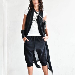May include: A woman wearing a black sleeveless cardigan over a white t-shirt with a skull graphic. She is wearing black pants with a drawstring waist and black and white sneakers.