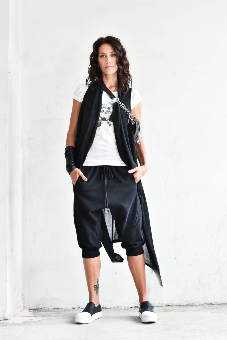 May include: A woman wearing a black sleeveless cardigan over a white t-shirt with a skull graphic. She is wearing black pants with a drawstring waist and black and white sneakers.