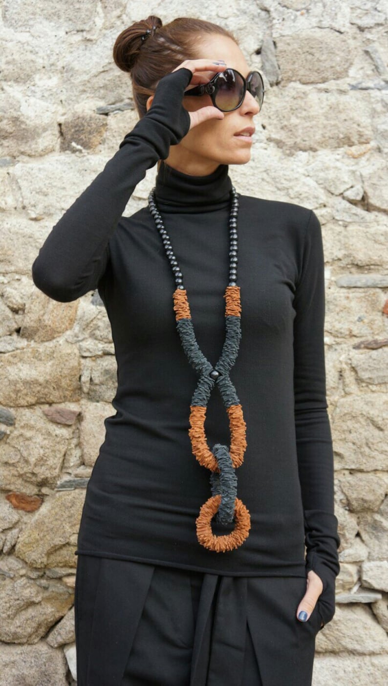 May include: A woman wearing a black turtleneck sweater and black pants with a long, brown and black necklace. She is wearing sunglasses and has her hair pulled back.