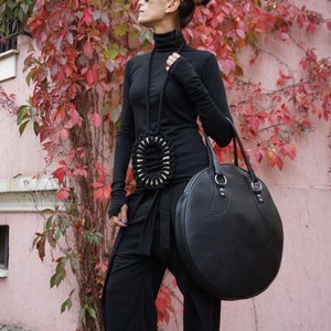 May include: A woman wearing a black turtleneck sweater, black pants, and white sneakers. She is carrying a large, round, black leather bag with a strap. She is standing in front of a wall covered in red ivy.