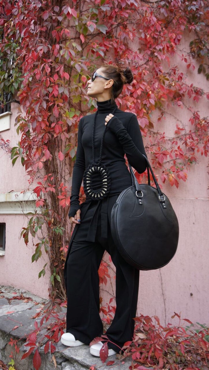 May include: A woman wearing a black turtleneck sweater, black pants, and white sneakers. She is carrying a large, round, black leather bag with a strap. She is standing in front of a wall covered in red ivy.