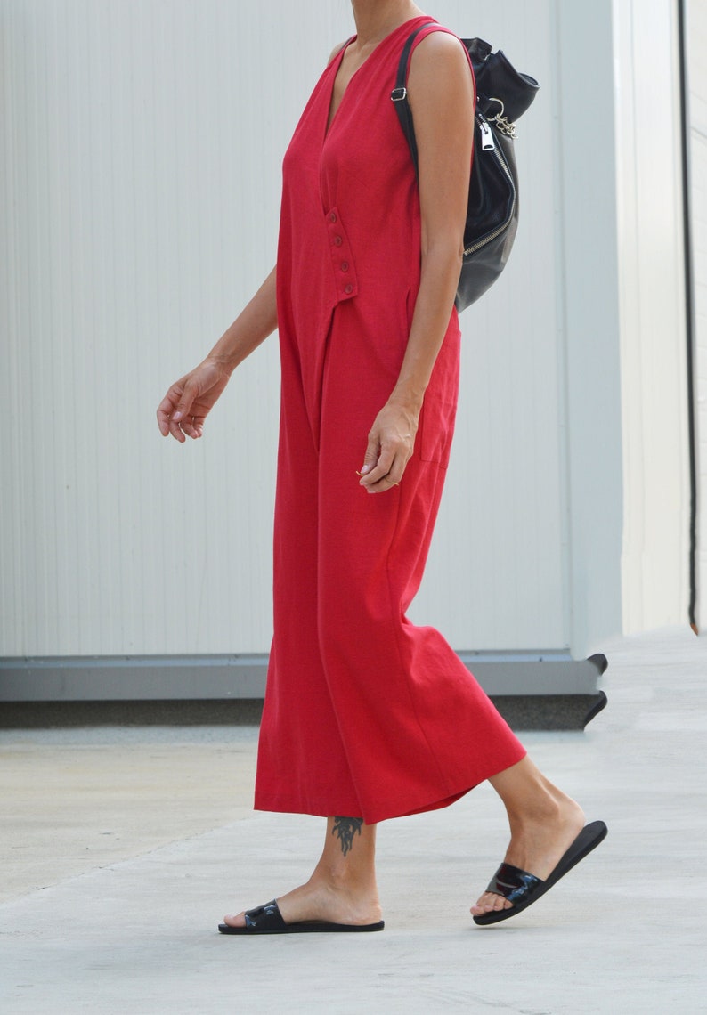 May include: A woman wearing a red sleeveless jumpsuit with a black backpack. She is wearing black sandals and walking on a concrete surface.