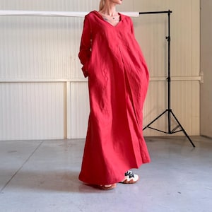 Può includere: Un lungo abito rosso, fluido, con maniche lunghe e scollo a V. L'abito ha una vestibilità ampia e tasche. La modella indossa sandali bianchi e neri. Lo sfondo è un muro bianco e un treppiede nero.