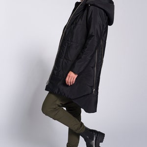 May include: A black hooded puffer jacket with a zipper down the back. The jacket is worn by a person wearing green pants and black boots.