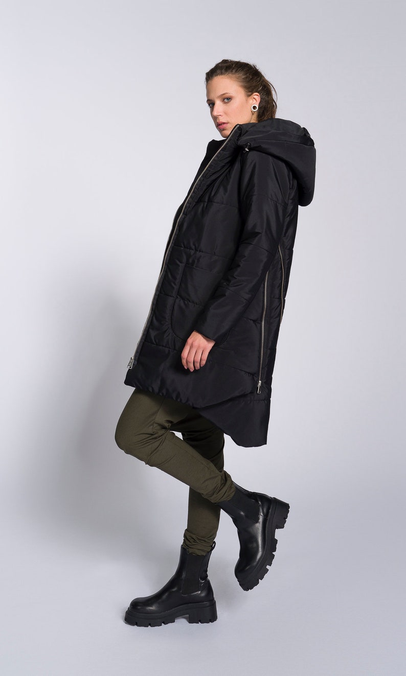 May include: A black hooded puffer jacket with a zipper down the back. The jacket is worn by a person wearing green pants and black boots.