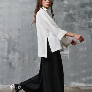 May include: A woman wearing a white linen shirt with a side slit and black wide-leg pants. She is wearing black and white high-top sneakers with red accents.