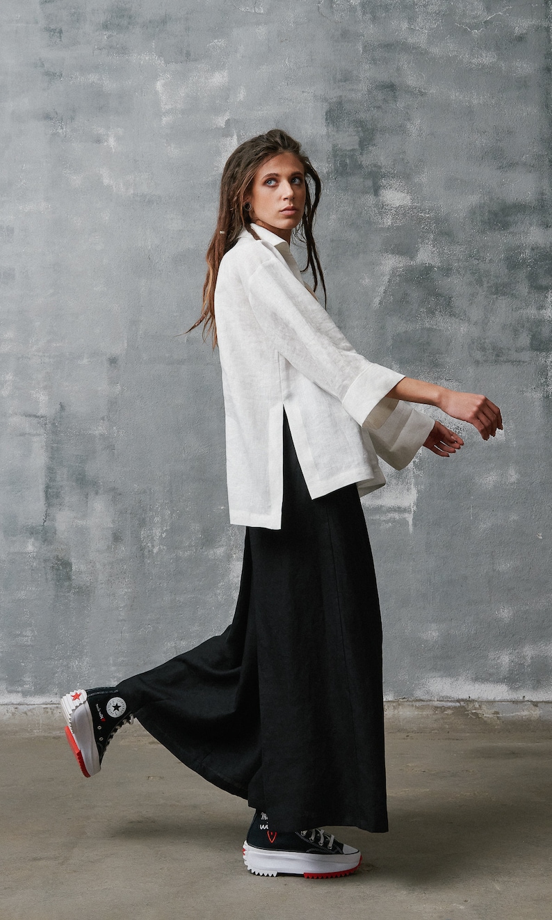 May include: A woman wearing a white linen shirt with a side slit and black wide-leg pants. She is wearing black and white high-top sneakers with red accents.