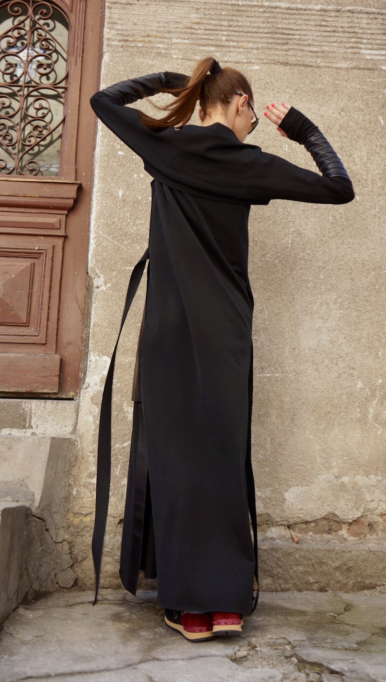 K&ouml;nnte beinhalten: Ein langes schwarzes Kleid mit langen &Auml;rmeln und einem Bindeg&uuml;rtel. Das Kleid hat einen Schlitz an der Seite und die &Auml;rmel haben Lederapplikationen.