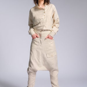 May include: A woman wearing a light beige linen jumpsuit with a long-sleeved shirt and pants. The pants have a unique, loose fit and a large pocket on the front. She is wearing white sneakers with a tan sole.