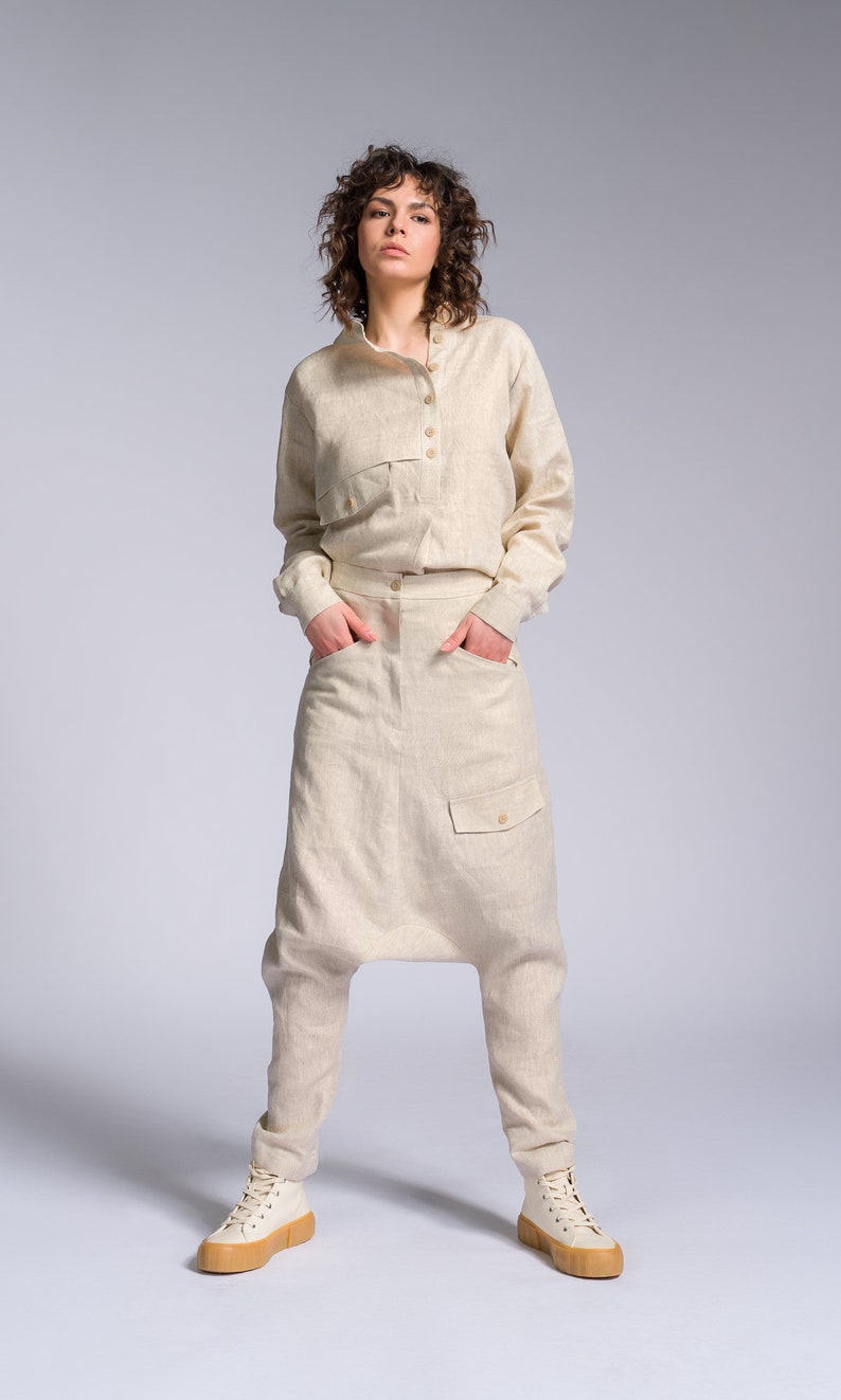May include: A woman wearing a light beige linen jumpsuit with a long-sleeved shirt and pants. The pants have a unique, loose fit and a large pocket on the front. She is wearing white sneakers with a tan sole.