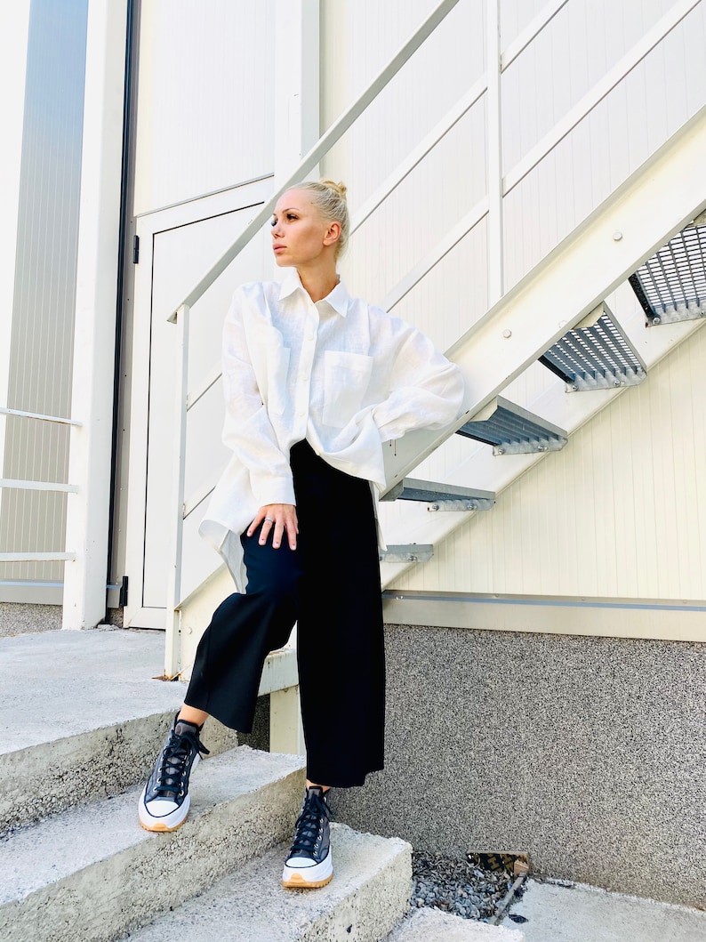 May include: A person wearing a white button-down shirt, black pants, and black high-top sneakers with a white sole and tan trim. The person is standing on concrete steps next to a metal staircase.