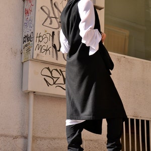 May include: A woman wearing a long black vest over a white shirt and black pants. She is wearing beige boots with a heart pattern. The woman is standing in front of a white wall with graffiti.