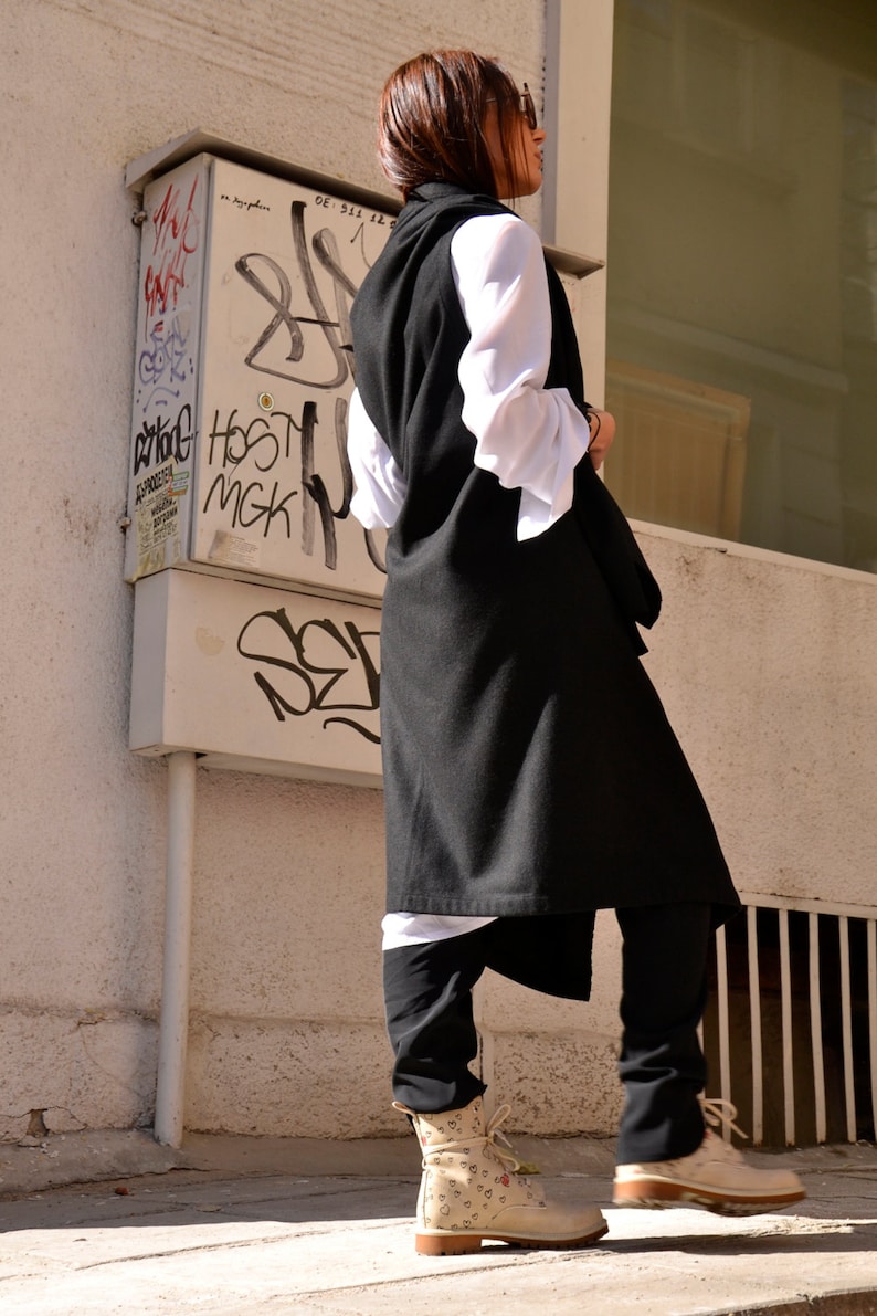 May include: A woman wearing a long black vest over a white shirt and black pants. She is wearing beige boots with a heart pattern. The woman is standing in front of a white wall with graffiti.