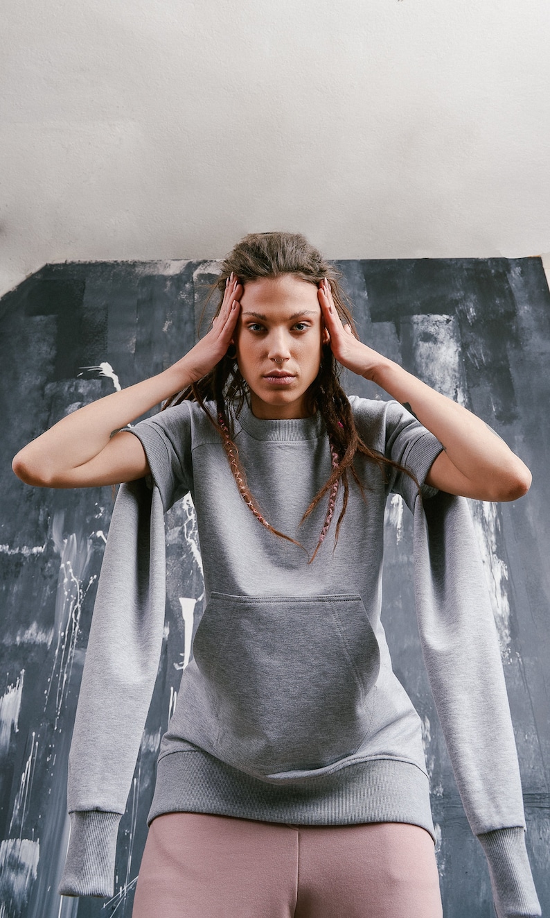 May include: A gray sweatshirt with long sleeves and a kangaroo pocket. The sleeves are extra long and have a wide, loose fit. The model is wearing the sweatshirt with pink pants.