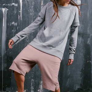 May include: A person wearing a gray sweatshirt with a large pocket and pink shorts. The sweatshirt has a double layer on the sleeves. The person is wearing white high-top sneakers.