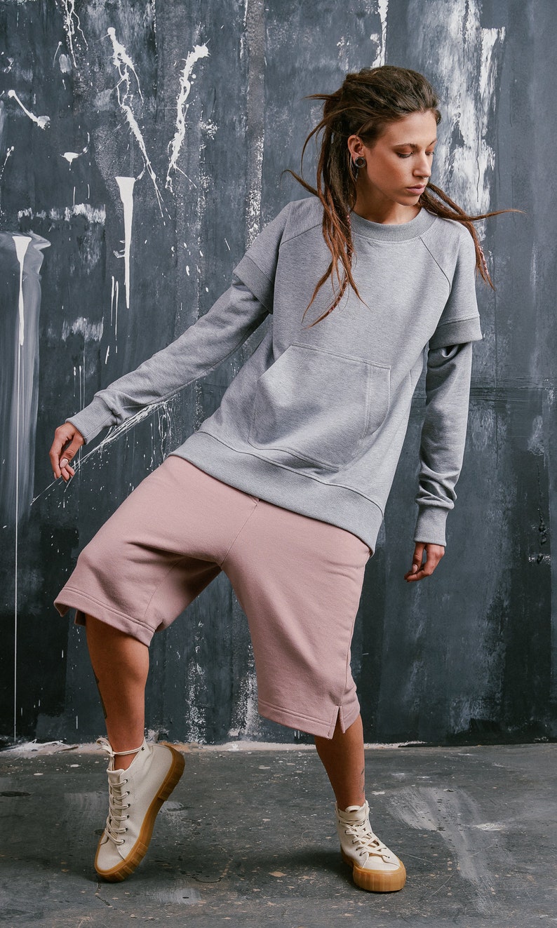 May include: A person wearing a gray sweatshirt with a large pocket and pink shorts. The sweatshirt has a double layer on the sleeves. The person is wearing white high-top sneakers.