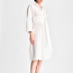 May include: A white linen shirt dress with a self-tie belt. The dress has a collared neckline and long sleeves.