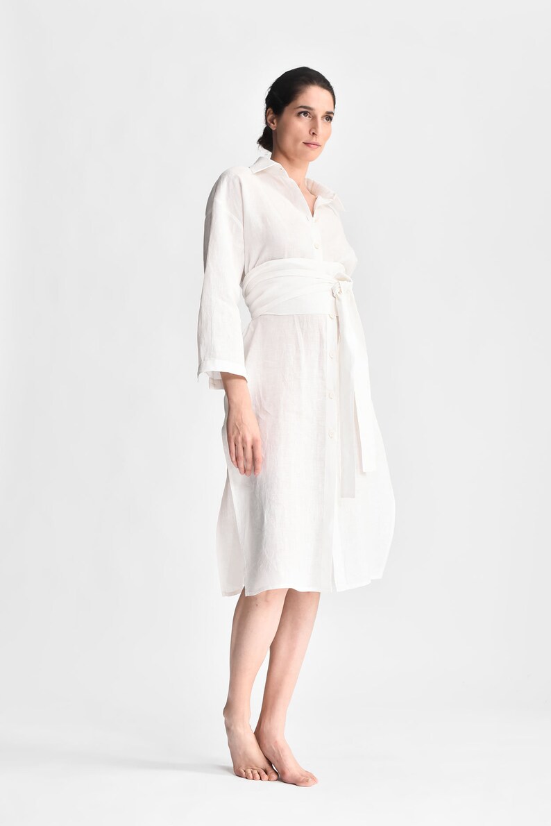 May include: A white linen shirt dress with a self-tie belt. The dress has a collared neckline and long sleeves.