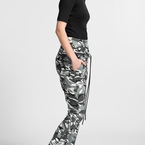 May include: A woman wearing a black cold shoulder top and camouflage pants with a drawstring. She is wearing black high-top sneakers with a white platform.