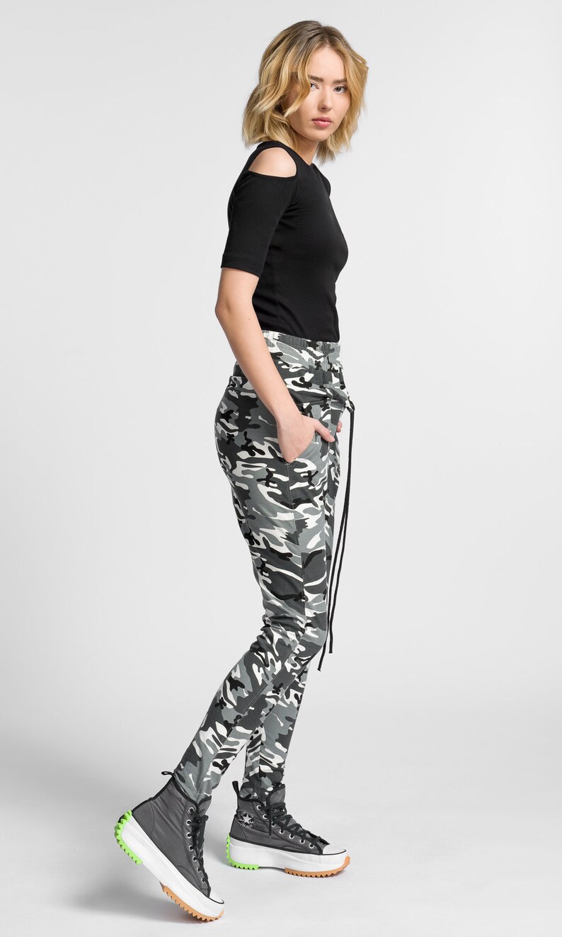 May include: A woman wearing a black cold shoulder top and camouflage pants with a drawstring. She is wearing black high-top sneakers with a white platform.