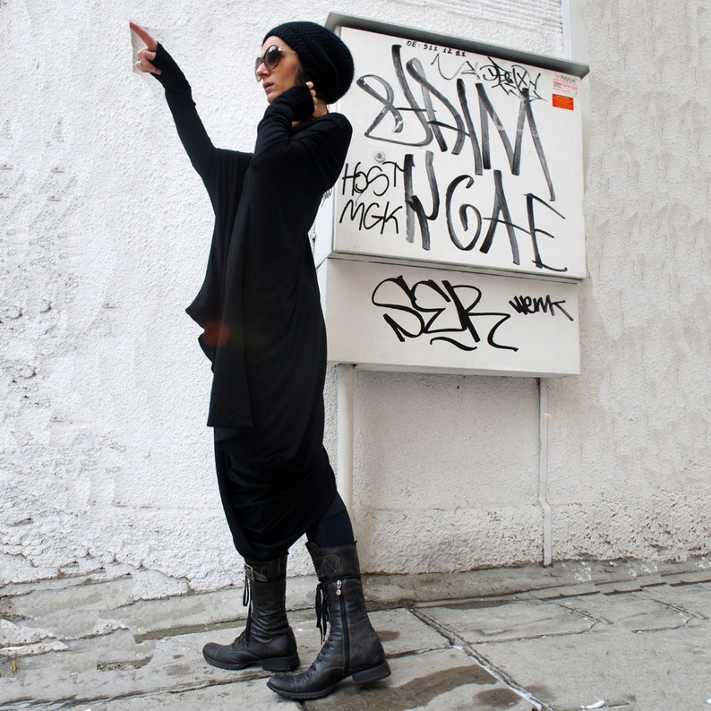 Op de afbeelding: Een vrouw draagt een zwarte lange mouwen tuniek met een zwarte beanie en zonnebril. Ze staat voor een witte muur met graffiti erop. De graffiti zegt "Brian McGee" en "SER".