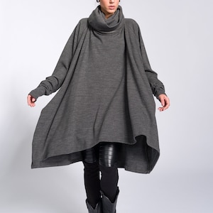 Könnte beinhalten: Ein grauer Oversize-Rollkragenpullover mit langen Ärmeln und einem High-Low-Saum. Der Pullover wird mit schwarzen Leggings und schwarzen Cowboy-Stiefeln getragen.