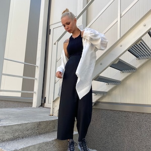 May include: A woman wearing a black tank top, black wide-leg pants, a white button-down shirt, and black sneakers with white soles and brown trim. She is standing on concrete steps next to a metal staircase.