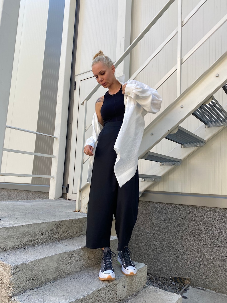 May include: A woman wearing a black tank top, black wide-leg pants, a white button-down shirt, and black sneakers with white soles and brown trim. She is standing on concrete steps next to a metal staircase.
