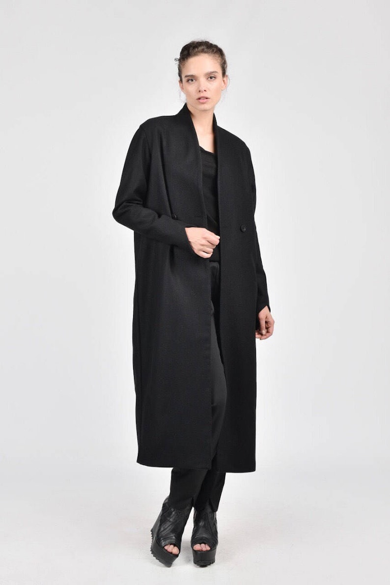 May include: A long black coat with a collar and double breasted closure. The coat is worn by a person with dark hair and is standing on a white background.