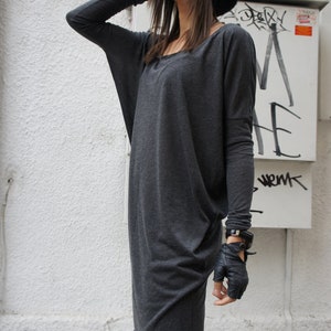 Puede incluir: Una mujer con una túnica gris de manga larga, un gorro negro y gafas de sol. También lleva guantes de cuero negro y pantalones negros.