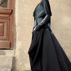 May include: A woman wearing a black long-sleeved top with a black skirt and black heeled sandals. She is standing in front of a wall with a door.