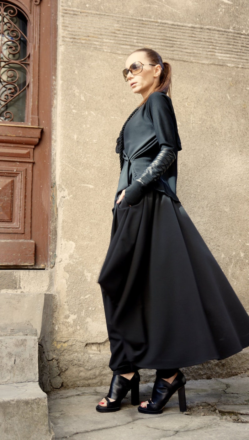 May include: A woman wearing a black long-sleeved top with a black skirt and black heeled sandals. She is standing in front of a wall with a door.