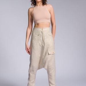 May include: A woman wearing a light brown tank top and light brown linen pants with a unique design. She is wearing white sneakers with a light brown sole.