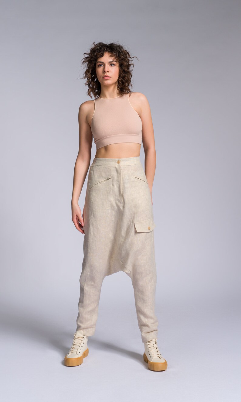May include: A woman wearing a light brown tank top and light brown linen pants with a unique design. She is wearing white sneakers with a light brown sole.