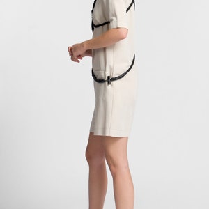 May include: A woman wearing a white linen jumpsuit with a black rope belt and black rope straps. She is wearing chunky trainers with black, white and neon yellow accents.