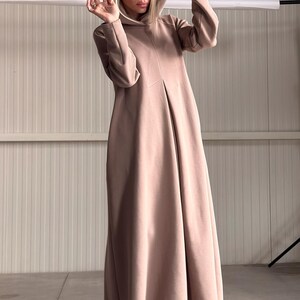 May include: A long, hooded dress in a soft, muted rose color. The dress features a flowing silhouette and a hood. The model is wearing the dress in a studio setting. The dress is made of a soft, comfortable material.