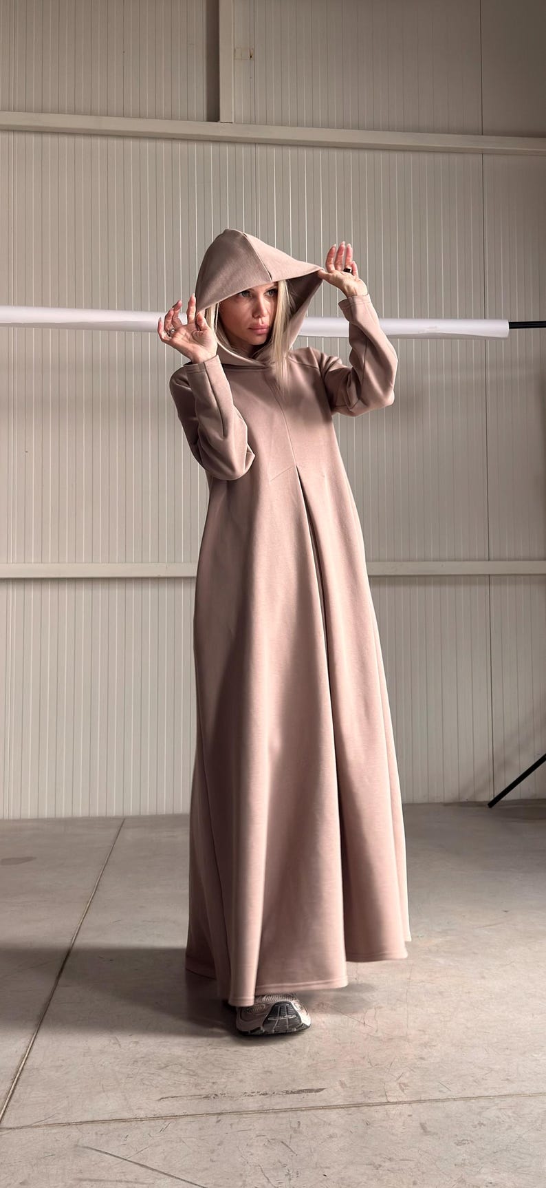 May include: A long, hooded dress in a soft, muted rose color. The dress features a flowing silhouette and a hood. The model is wearing the dress in a studio setting. The dress is made of a soft, comfortable material.