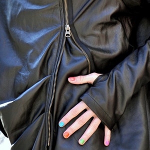 May include: A black leather jacket with a zipper closure. The jacket is unzipped and the person's hand is visible with colorful nail polish.