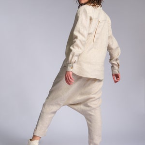 May include: A woman wearing a beige linen shirt and matching pants. She is wearing white sneakers with a brown sole. The woman is standing on a white background and is looking over her shoulder.