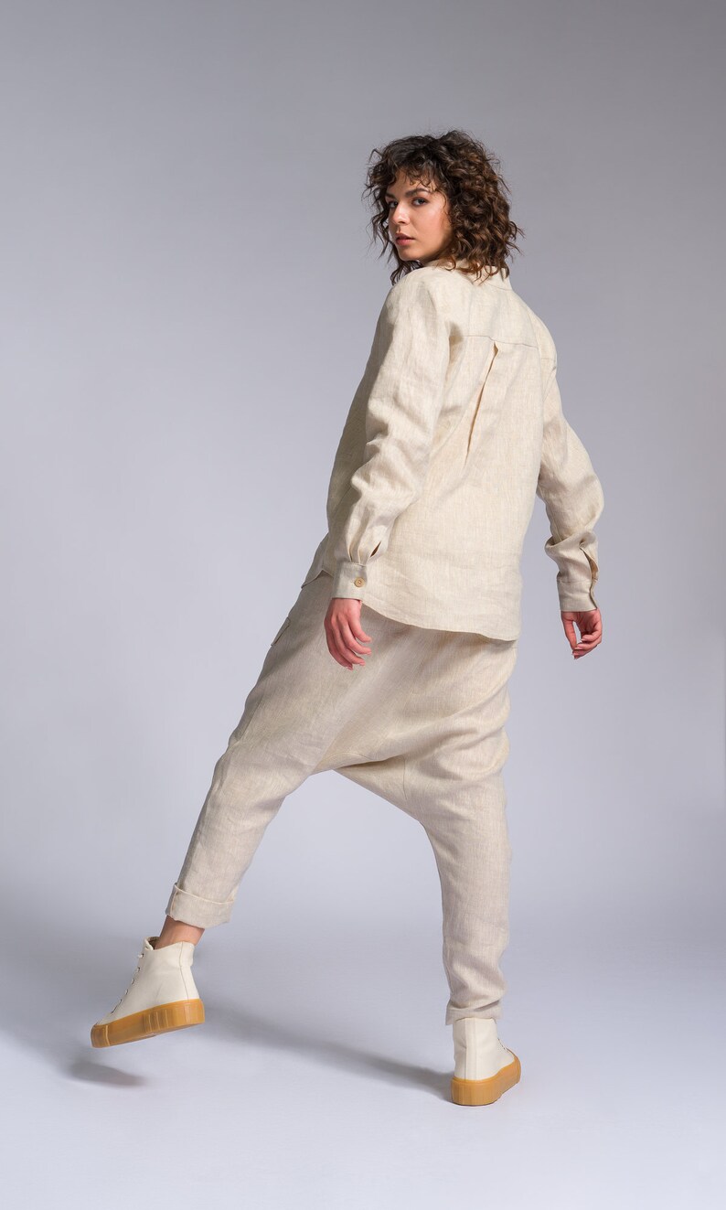 May include: A woman wearing a beige linen shirt and matching pants. She is wearing white sneakers with a brown sole. The woman is standing on a white background and is looking over her shoulder.