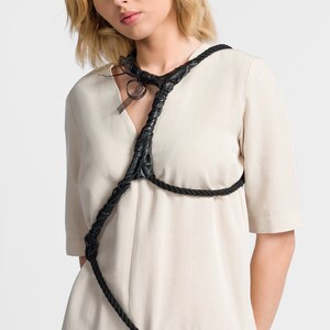 May include: A woman wearing a beige linen dress with a black leather rope detail across the chest and shoulders.