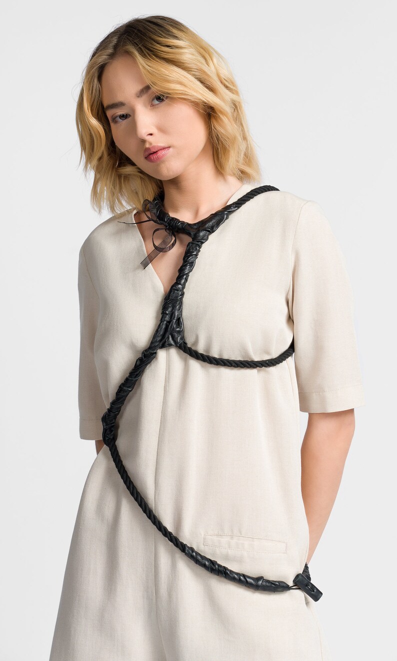 May include: A woman wearing a beige linen dress with a black leather rope detail across the chest and shoulders.