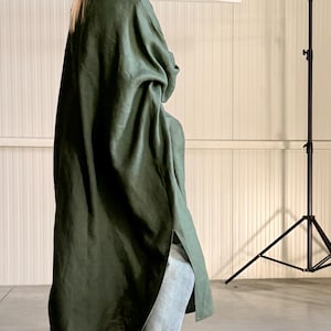 Pode incluir: Um casaco comprido de linho verde-oliva com uma fenda lateral, usado sobre jeans azul claro e tênis brancos. O casaco tem um corte folgado e uma gola. O fundo é um estúdio neutro.