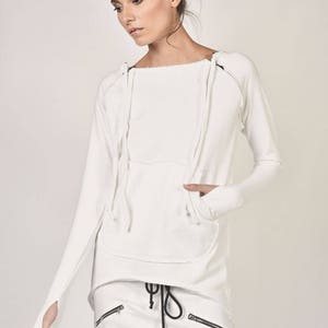 Sudadera asimétrica de algodón: color blanco hueso, con orificios para los pulgares, detalle de cremallera en los hombros y bolsillo tipo canguro frontal.