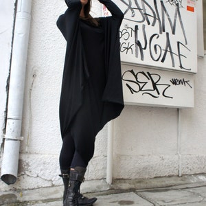 Op de afbeelding: Een vrouw draagt een zwarte lange mouwen tuniek met een cowl hals en zwarte leggings. Ze draagt een zonnebril en zwarte laarzen. De achtergrond is een witte muur met graffiti.